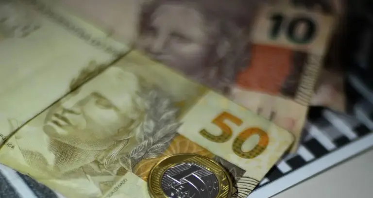 A imagem em close-up apresenta uma composição de cédulas e uma moeda do Real brasileiro, com um foco seletivo que destaca a nota de R50eumamoedadeR1. Em primeiro plano e com maior nitidez, está uma nota de cinquenta reais (R50),comon u ˊ mero"50"emdestaqueeaef ı ˊ giedaRep u ˊ blica.Parcialmentesobrepostaaela,naparteinferiordireita,h a ˊ umamoedadeumreal(R1), que reflete a luz. Ao fundo, e com um desfoque que suaviza seus detalhes, é possível identificar parcialmente outras cédulas. Uma delas, na parte superior direita, parece ser uma nota de dez reais (R$10), com o número "10" visível. Outra nota está mais à esquerda, também com a efígie da República. O fundo é escuro e não permite identificar o ambiente. A iluminação da imagem é suave, mas com alguns pontos de brilho na moeda e nas superfícies das notas, sugerindo uma fonte de luz indireta. A composição geral evoca o tema de finanças, economia ou transações monetárias.