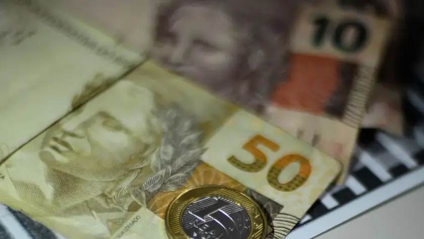 A imagem em close-up apresenta uma composição de cédulas e uma moeda do Real brasileiro, com um foco seletivo que destaca a nota de R50eumamoedadeR1. Em primeiro plano e com maior nitidez, está uma nota de cinquenta reais (R50),comon u ˊ mero"50"emdestaqueeaef ı ˊ giedaRep u ˊ blica.Parcialmentesobrepostaaela,naparteinferiordireita,h a ˊ umamoedadeumreal(R1), que reflete a luz. Ao fundo, e com um desfoque que suaviza seus detalhes, é possível identificar parcialmente outras cédulas. Uma delas, na parte superior direita, parece ser uma nota de dez reais (R$10), com o número "10" visível. Outra nota está mais à esquerda, também com a efígie da República. O fundo é escuro e não permite identificar o ambiente. A iluminação da imagem é suave, mas com alguns pontos de brilho na moeda e nas superfícies das notas, sugerindo uma fonte de luz indireta. A composição geral evoca o tema de finanças, economia ou transações monetárias.