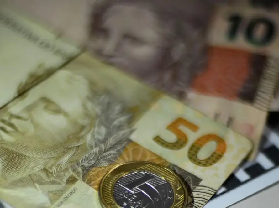 A imagem em close-up apresenta uma composição de cédulas e uma moeda do Real brasileiro, com um foco seletivo que destaca a nota de R50eumamoedadeR1. Em primeiro plano e com maior nitidez, está uma nota de cinquenta reais (R50),comon u ˊ mero"50"emdestaqueeaef ı ˊ giedaRep u ˊ blica.Parcialmentesobrepostaaela,naparteinferiordireita,h a ˊ umamoedadeumreal(R1), que reflete a luz. Ao fundo, e com um desfoque que suaviza seus detalhes, é possível identificar parcialmente outras cédulas. Uma delas, na parte superior direita, parece ser uma nota de dez reais (R$10), com o número "10" visível. Outra nota está mais à esquerda, também com a efígie da República. O fundo é escuro e não permite identificar o ambiente. A iluminação da imagem é suave, mas com alguns pontos de brilho na moeda e nas superfícies das notas, sugerindo uma fonte de luz indireta. A composição geral evoca o tema de finanças, economia ou transações monetárias.
