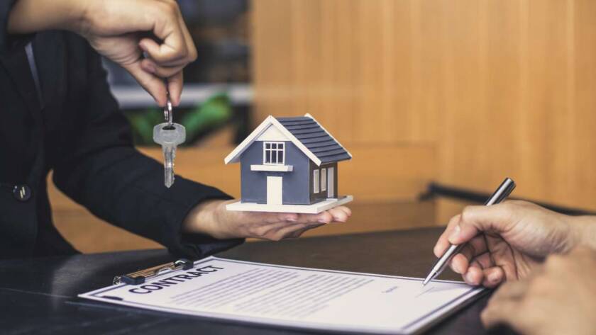 A imagem retrata uma transação imobiliária ou um acordo relacionado à compra/venda de uma propriedade, com foco em elementos simbólicos e documentais. No centro da imagem, uma mão (provavelmente de um agente imobiliário ou vendedor) segura uma pequena maquete de uma casa moderna, com telhado escuro e paredes claras, representando a propriedade em questão. Acima da maquete, a mesma mão (ou uma segunda mão próxima) estende um chaveiro com uma única chave prateada, simbolizando a posse da casa. Na parte inferior direita da imagem, outra mão está em processo de assinar um documento, que está em uma prancheta preta. A palavra "CONTRATO" (ou "CONTRACT") é claramente visível no topo do documento. A caneta esferográfica preta na mão indica a finalização ou formalização do acordo. O fundo é um ambiente interno, possivelmente um escritório ou mesa de negociação, com uma parede de madeira clara ao fundo, transmitindo uma atmosfera profissional e séria. A iluminação é suave e uniforme, destacando os elementos principais da cena. A imagem comunica os conceitos de negociação, propriedade, acordo legal e a entrega de chaves em um contexto imobiliário.