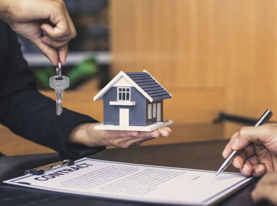 A imagem retrata uma transação imobiliária ou um acordo relacionado à compra/venda de uma propriedade, com foco em elementos simbólicos e documentais. No centro da imagem, uma mão (provavelmente de um agente imobiliário ou vendedor) segura uma pequena maquete de uma casa moderna, com telhado escuro e paredes claras, representando a propriedade em questão. Acima da maquete, a mesma mão (ou uma segunda mão próxima) estende um chaveiro com uma única chave prateada, simbolizando a posse da casa. Na parte inferior direita da imagem, outra mão está em processo de assinar um documento, que está em uma prancheta preta. A palavra "CONTRATO" (ou "CONTRACT") é claramente visível no topo do documento. A caneta esferográfica preta na mão indica a finalização ou formalização do acordo. O fundo é um ambiente interno, possivelmente um escritório ou mesa de negociação, com uma parede de madeira clara ao fundo, transmitindo uma atmosfera profissional e séria. A iluminação é suave e uniforme, destacando os elementos principais da cena. A imagem comunica os conceitos de negociação, propriedade, acordo legal e a entrega de chaves em um contexto imobiliário.