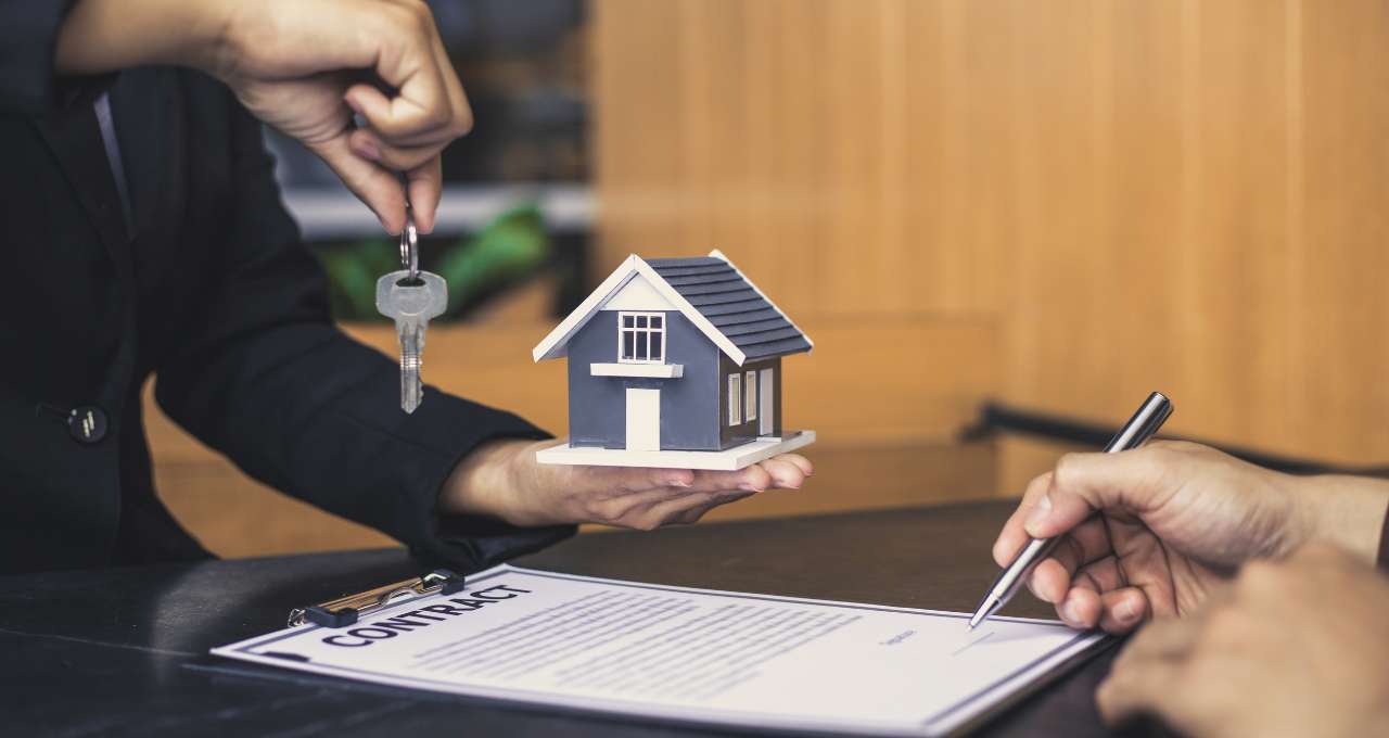 A imagem retrata uma transação imobiliária ou um acordo relacionado à compra/venda de uma propriedade, com foco em elementos simbólicos e documentais. No centro da imagem, uma mão (provavelmente de um agente imobiliário ou vendedor) segura uma pequena maquete de uma casa moderna, com telhado escuro e paredes claras, representando a propriedade em questão. Acima da maquete, a mesma mão (ou uma segunda mão próxima) estende um chaveiro com uma única chave prateada, simbolizando a posse da casa. Na parte inferior direita da imagem, outra mão está em processo de assinar um documento, que está em uma prancheta preta. A palavra "CONTRATO" (ou "CONTRACT") é claramente visível no topo do documento. A caneta esferográfica preta na mão indica a finalização ou formalização do acordo. O fundo é um ambiente interno, possivelmente um escritório ou mesa de negociação, com uma parede de madeira clara ao fundo, transmitindo uma atmosfera profissional e séria. A iluminação é suave e uniforme, destacando os elementos principais da cena. A imagem comunica os conceitos de negociação, propriedade, acordo legal e a entrega de chaves em um contexto imobiliário.